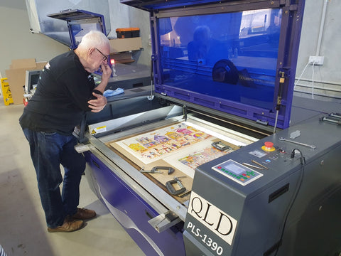 A photo of a man standing over a machine that appears to be a laser cutter, preparing for a puzzle to be newly cut