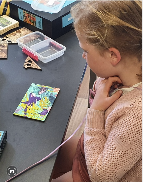 A photo of a young girl, with blonde hair, wearing a pink sweater sitting at a table, looking at a small miniature puzzle which has been freshly assembled.