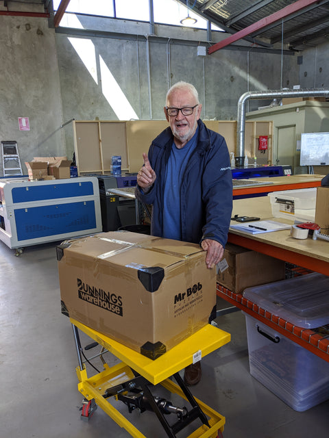 A photo of a man standing over a cardboard box, pointing at the ceiling. The cardboard box has a Bunnings Warehouse logo on it.