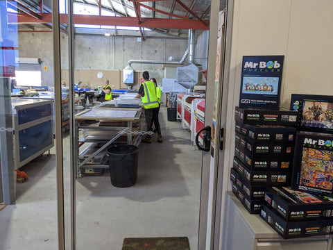 An internal photo of the Mr Bob Puzzles factory with various tables and workers in hi-vis clothing around. There are various Mr Bob Puzzles puzzles stacked on top of each other in front of the doorway.